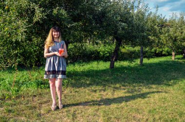 Gri elbiseli güzel bir kadın, güneşli bir günde serinletici soğuk bir içeceğin tadını çıkarırken yemyeşil bir meyve bahçesinde duruyor, doğanın sükunetini kucaklıyor.