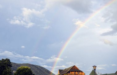 Bulutlu bir gökkuşağına karşı canlı bir gökkuşağının yer aldığı, kırsal binalar ve yemyeşil verimli bir köy manzarası. Yağmurdan sonra kırsal yaşamın sakinliğini ve güzelliğini yakalar.