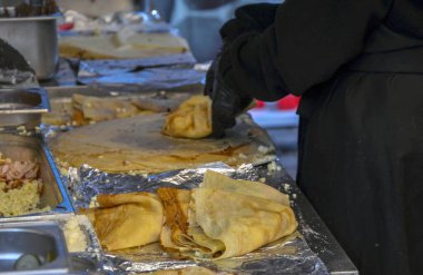 Bir sokak yemeği satıcısında yapılan kreplerin yakın çekim görüntüsü. Görüntü katlanmış krepleri, malzemeleri ve aşçının ellerini bir araya topluyor.