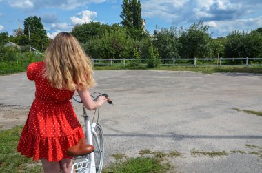 Kırmızı benekli elbiseli bir kadının arkası huzurlu bir kırsal alanda bisiklet sürüyor. Sahne, güneşli bir günde doğayla bir özgürlük, huzur ve bağ hissi uyandırıyor.