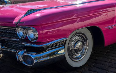 Canlı pembe klasik Cadillac Eldorado arabası parlak krom detaylar ve güneşli bir kaldırım taşı caddesinde park edilmiş büyük beyaz duvar lastikleriyle sergileniyor.