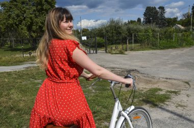 Kırmızı benekli elbiseli neşeli kadın, yeşil ve açık gökyüzü ile çevrili sakin bir manzarada bisikletin üzerinde oturuyor, doğada aktif ve neşeli bir anın tadını çıkarıyor.