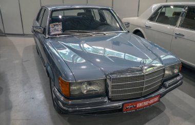 Görüntü gümüş bir Mercedes-Benz 450SE gösteriyor, klasik lüks bir sedan. Araba diğer klasik araçlarla birlikte bir galeride park halinde.