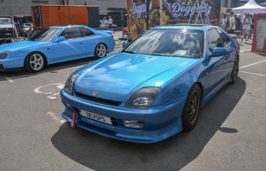 Pırıltılı mavi 1997 Honda Prelude, 1990 'lardan kalma popüler bir Japon spor coupe açık hava etkinliğinde park etmiş, canlı rengi ve modern tasarımı ile dikkat çekiyor.