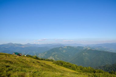 Huzurlu dağ manzarası, arka planda tepeleri ve tepeleri yuvarlanan yeşil bir ormanın yanında güzel mavi bir gökyüzünün altında güneş ışığıyla yıkanmış bir ev gösteriyor. Karpatlar, Ukrayna