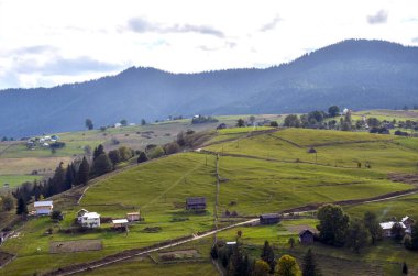 Yeşillik yeşillik ve bulutlu bir gökyüzü altında uzak ormanlık dağlarla çevrili, küçük evler ve çiftlik yapılarıyla görülen yeşil yuvarlanan tepelerin olduğu ideolojik kırsal bir manzara. Karpatlar, Ukrayna