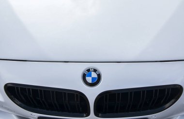 Beyaz arabanın kaputunda gösterilen ikonik BMW logosunun ayrıntılı görünümü, markanın basit ve temiz kompozisyon içindeki lüks ve tasarım üzerine vurgusunu vurguluyor