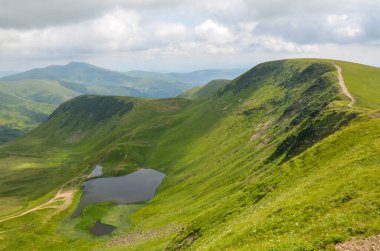 Güzel yeşil tepeler bulutlu bir gökyüzünün altında sakin bir Dogyaska gölünü çevreleyerek doğa severler, yürüyüş ve fotoğrafçılık için mükemmel bir manzara yaratır. Svydovets, Karpatlar, Ukrayna