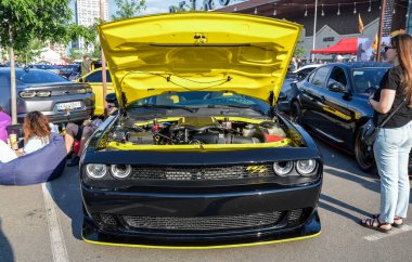 Parlak siyah ve sarı Dodge Challenger kaputu açık, güçlü motorunu bir araba fuarında sergiliyor kaslı arabanın tasarımını ve ikonik durumunu vurguluyor. 
