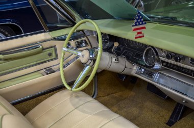 Açık yeşil direksiyonu ve krom aksanlı gösterge paneli olan klasik bir Cadillac DeVille arabasının içi. İç kısımda bej renkli deri koltuklar ve halı kaplı zemin var.