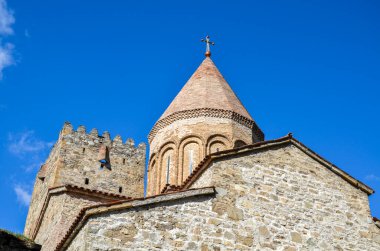 Crenellasyonlu taş kule, açık mavi gökyüzüne karşı konik tuğla bir kubbe ve altın bir haçın bulunduğu bir kilisenin yanında duruyor. Georgia 'daki Ananuri Kalesi Kompleksi