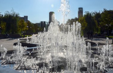Şehir parkında, arka planda kentsel binalar bulunan açık ve güneşli gökyüzünün altında çeşitli şekillerde su püskürten büyük bir çeşme. Erivan, Ermenistan