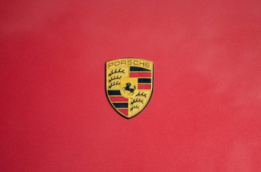 Porsche ambleminin düz, koyu kırmızı arkaplana yakın görüntüsü. Amblem ortalanmıştır ve sarı bir alanda siyah atlı ikonik Porsche armasını içerir.