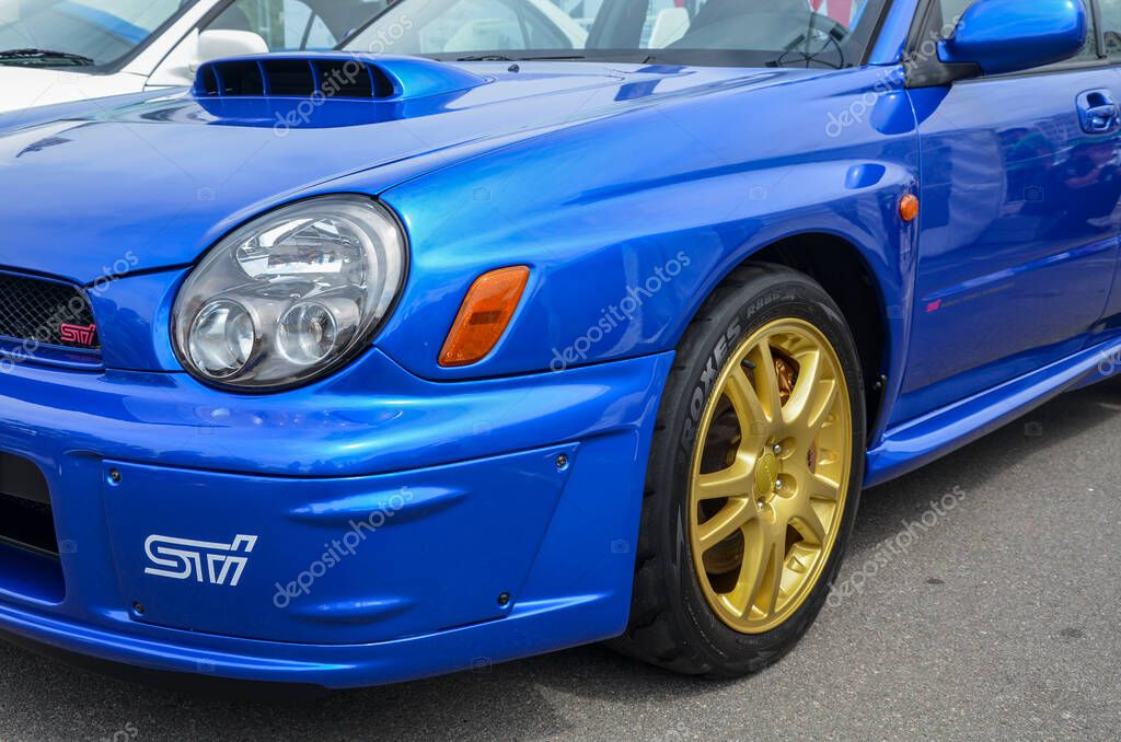 Close-up shot of the front of a blue Subaru Impreza WRX STI. The car features a distinctive hood scoop, gold-colored wheels, and the 
