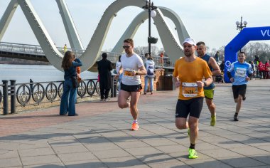 Bir maraton yarışında nehir kenarında koşan enerjik katılımcılar, takım çalışması, çaba ve şehir hayatı sergiliyorlar.
