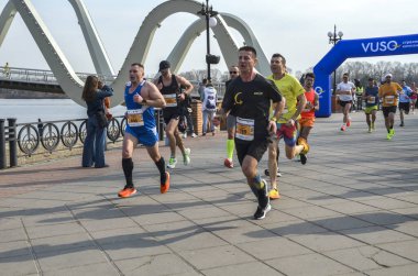 Bir maraton yarışında nehir kenarında koşan enerjik katılımcılar, takım çalışması, çaba ve şehir hayatı sergiliyorlar.