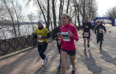 Atletik kıyafetler giymiş bir grup koşucu nehir kenarında ağaçlar ve sakin nehirlerle koşuyor. Güneşli bir günde açık hava fitness ve grup katılımı