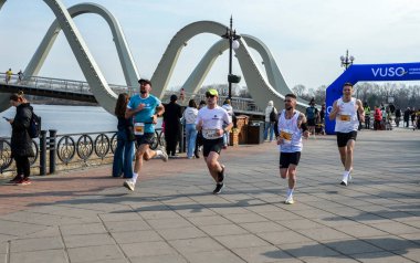 Bir maraton yarışında nehir kenarında koşan enerjik katılımcılar, takım çalışması, çaba ve şehir hayatı sergiliyorlar.