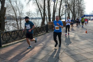 Nehir yolu boyunca koşan bir maratona katılanlar güneşli bir günde açık hava ortamında spor, takım çalışması ve kararlılık gösteriyorlar.