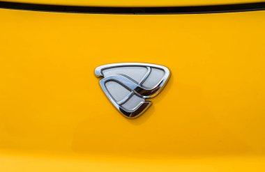 Sarı bir arabanın kaputunda gümüş renkli Mazda araba amblemi. Amblem kıvrımlı çizgileri olan stilize bir kalkanı andırıyor, markanın belirgin kimliğini vurguluyor.