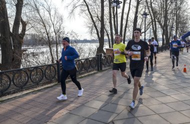 Bir grup koşucu manzaralı bir park yolu boyunca bir maratona katılıyor. Bireyler enerjik ve odaklanmış görünüyor, fiziksel aktiviteden zevk alıyorlar.