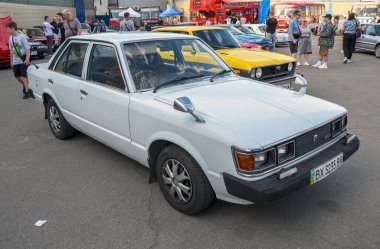 Beyaz Toyota Carina, krom yan aynaları, siyah tamponları ve özgün ön ızgarası ile öne çıkıyor, retro otomotiv estetiğini somutlaştırıyor.