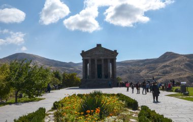 Antik Garni tapınağı, parçalı bulutlu bir gökyüzünün altındaki dağlara bakan bir tepede duruyor. Ziyaretçiler tarihi anıtı takdir ediyorlar