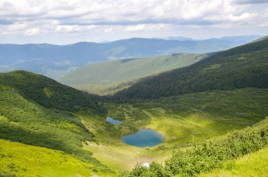 Etrafı yemyeşil orman dağlarıyla çevrili tenha bir vadide bulutlu bir gökyüzünün altında sakin mavi Vorozheska gölü bulunuyor. Svydovets sırtı, Karpatlar, Ukrayna 