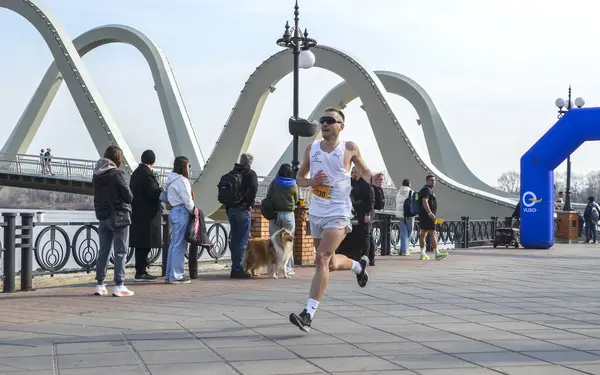 Modern bir köprünün yakınında izleyiciler tarafından çevrelenmiş rekabetçi bir yarışmada koşan atletik bir adam.