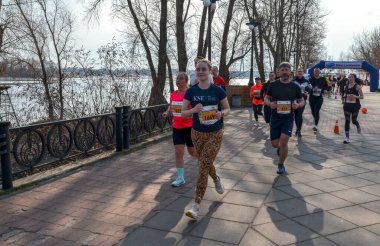 Koşucular canlı bir açık hava yarışına aktif olarak katılıyorlar. Nehir manzaralı ve ağaçlarla çevrili. 