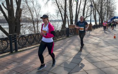 Koşucular canlı bir açık hava yarışına aktif olarak katılıyorlar. Nehir manzaralı ve ağaçlarla çevrili. 
