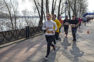 Koşucular canlı bir açık hava yarışına aktif olarak katılıyorlar. Nehir manzaralı ve ağaçlarla çevrili. 