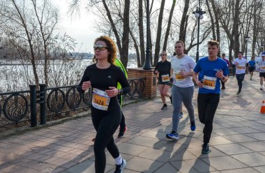 Bir grup atletizm sporcusu nehir kenarındaki patika boyunca maratona katılıyor. Koşucular, barışçıl bir günde açık hava fitness etkinliği sırasında kendilerini adamış ve enerjik görünüyorlar.