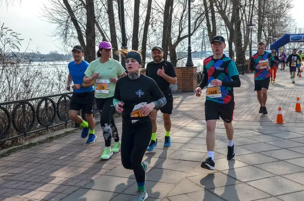 Bir grup atletizm sporcusu nehir kenarındaki patika boyunca maratona katılıyor. Koşucular, barışçıl bir günde açık hava fitness etkinliği sırasında kendilerini adamış ve enerjik görünüyorlar.