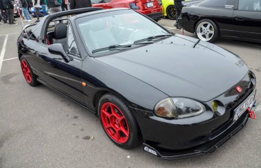 Parlak siyah Honda Del Sol, kırmızı tekerlekleriyle bir açık hava araba etkinliğinde, targa bluzunu sergiliyor. Arka planda diğer arabalar ve insanlar görünüyor.