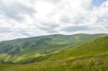Parçalı bulutlu bir gökyüzünün altında yuvarlanan yeşil tepelerin ve dağların huzurlu manzarası. Doğanın güzelliğinin ve huzurunun özünü yakalıyor. Karpat Dağları, Ukrayna