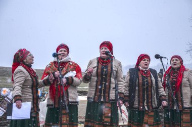 Renkli giysiler içinde bir grup geleneksel folk şarkıcısı dışarıda karlı zemine karşı gösteri yapıyor. Kültürel birlik, kutlama ve ahenk duyguları uyandırıyor.