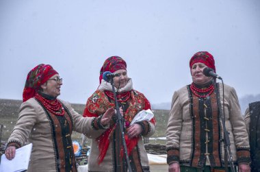 Renkli giysiler içinde bir grup geleneksel folk şarkıcısı dışarıda karlı zemine karşı gösteri yapıyor. Kültürel birlik, kutlama ve ahenk duyguları uyandırıyor.