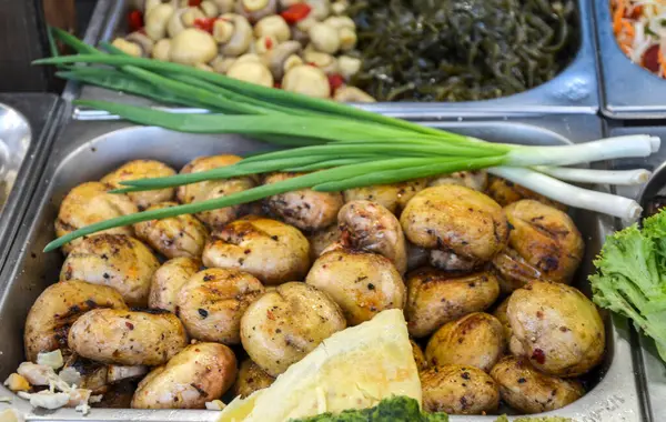 Açık büfe ortamında taze soğanla sunulan ızgara mantarların yakın çekim görüntüsü diğer iştah açıcı yemeklerle çevrili lezzetli ve sağlıklı bir mutfak seçeneği sunuyor.