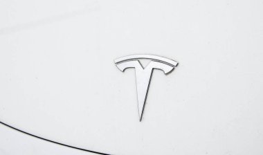 Yakın çekim beyaz bir araba yüzeyindeki gümüş Tesla logosunu gösteriyor. Görüntü biraz açılı, ikonik 