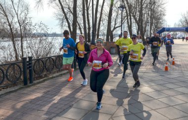 Bir grup koşucu, doğa tarafından çevrelenmiş nehir kenarındaki yürüyüş yolu boyunca düzenlenmiş bir açık hava etkinliğine katılıyor.