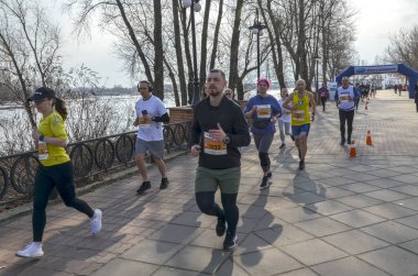 Bir grup koşucu, doğa tarafından çevrelenmiş nehir kenarındaki yürüyüş yolu boyunca düzenlenmiş bir açık hava etkinliğine katılıyor.