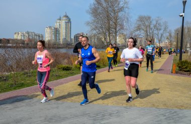 Bir grup sporcu, gündüz düzenlenen bir maraton sırasında nehir kenarında manzaralı bir patika boyunca koşuyor.