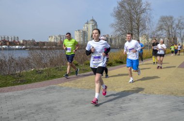Bir grup sporcu, gündüz düzenlenen bir maraton sırasında nehir kenarında manzaralı bir patika boyunca koşuyor.