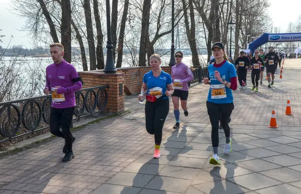 Bir grup koşucu, doğa tarafından çevrelenmiş nehir kenarındaki yürüyüş yolu boyunca düzenlenmiş bir açık hava etkinliğine katılıyor.
