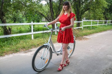 Kırmızı benekli elbise giyen bir kadın, sakin kırsal bir yolda bisikletle duruyor, yemyeşil, rahatlama, hareketlilik ve açık hava keyfi hissi yayıyor.