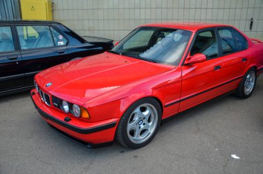 Muhteşem kırmızı klasik BMW E34 M5, ikonik spor sedan tasarımı sergiliyor. Arabada gümüş alaşımlı tekerlekler ve siyah süslemeli aksanlar var.