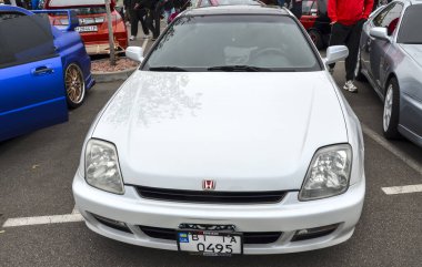 Parlak beyaz beşinci nesil Honda Prelude altın renkli alaşım tekerlekleri, parlak, düşük çizgili profili ve araba buluşmasında keskin farları var.
