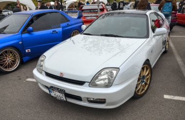 Parlak beyaz beşinci nesil Honda Prelude altın renkli alaşım tekerlekleri, parlak, düşük çizgili profili ve araba buluşmasında keskin farları var.