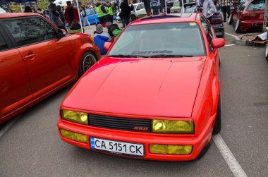 Canlı kırmızı Volkswagen Corrado 1990, sarı farlar ve düşük süspansiyonlu açık hava otomobil etkinliğinde gösteriliyor.